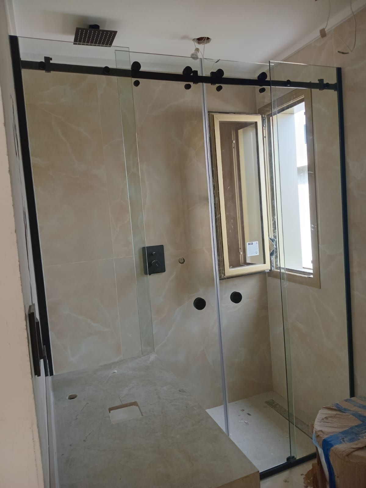 Mampara de ducha de cristal con marco negro, incluye puerta corredera, junto a una ventana en un baño.