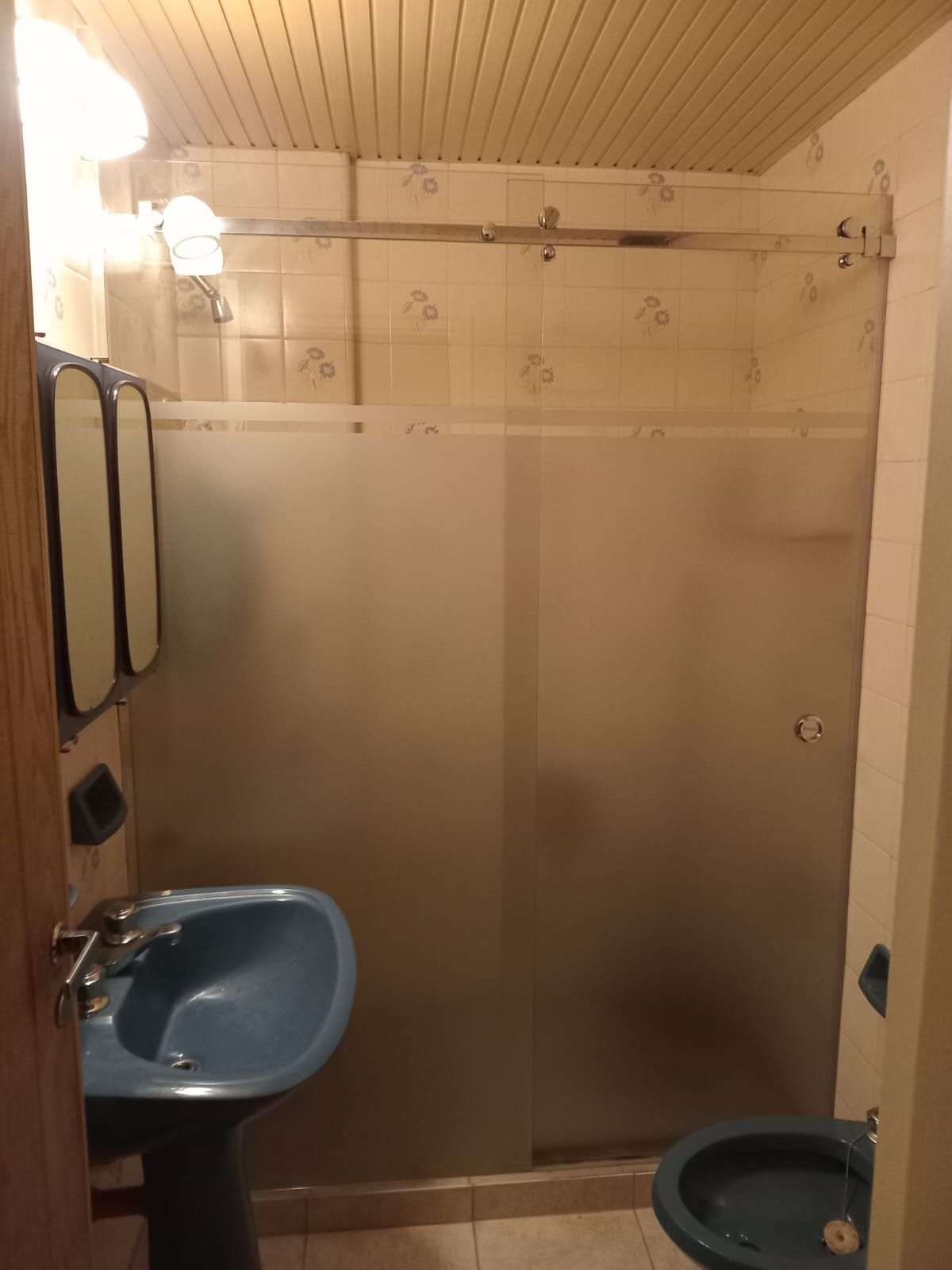 Baño con lavabo e inodoro azules, ducha con cristal esmerilado y botiquín con espejo.