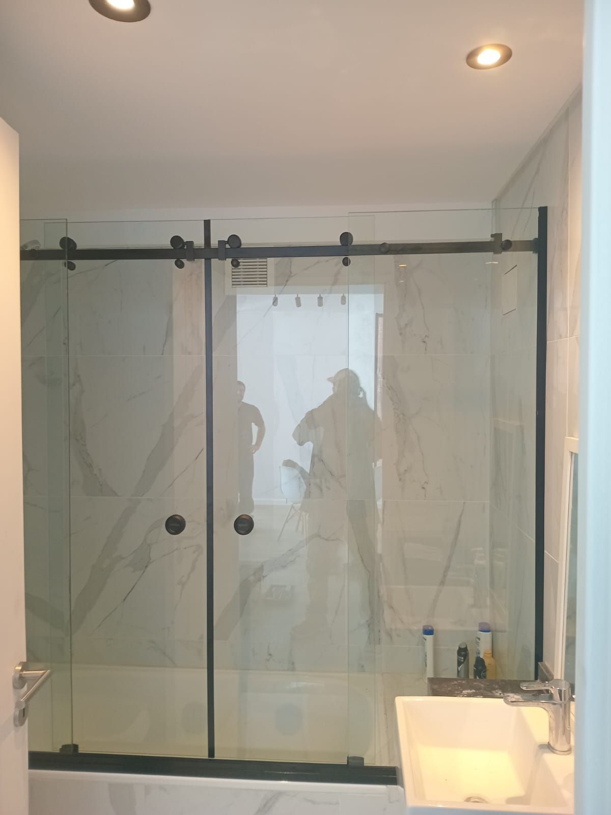 Ducha con puertas corredizas de cristal y herrajes negros. Una persona se refleja en el cristal. Azulejos blancos