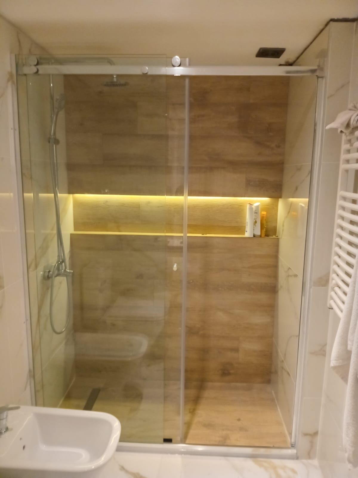 Ducha con puertas de cristal, pared de azulejos imitación madera con estante empotrado