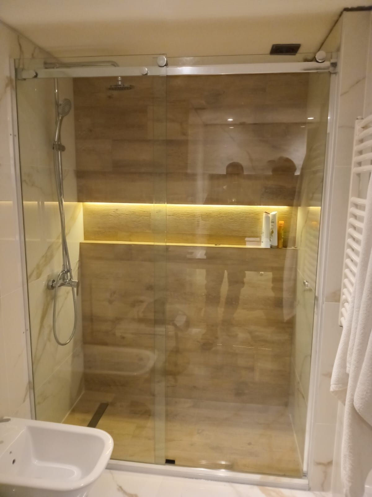 Ducha con puerta corrediza de cristal, revestimiento de madera y estante empotrado iluminado por una luz cálida.