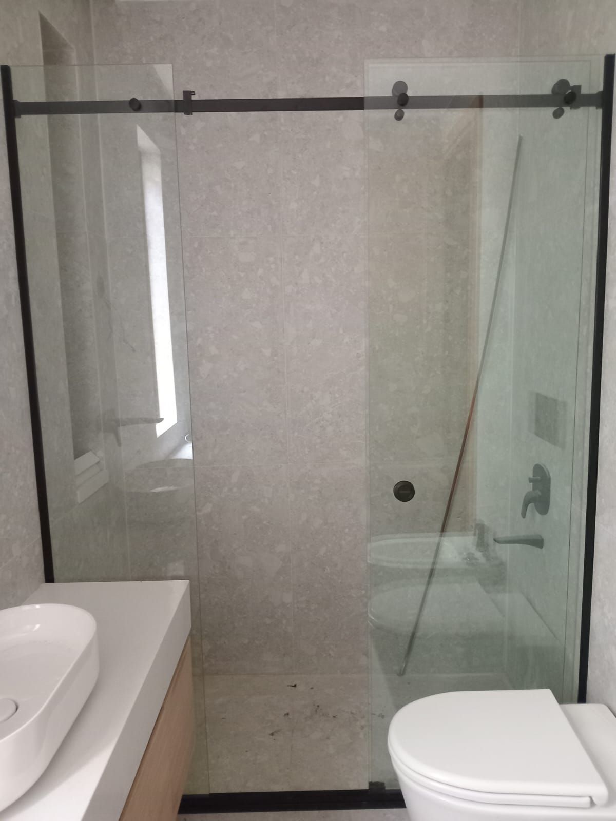 Baño con puerta de ducha de vidrio, marco negro, inodoro blanco y lavabo contra una pared gris texturizada.