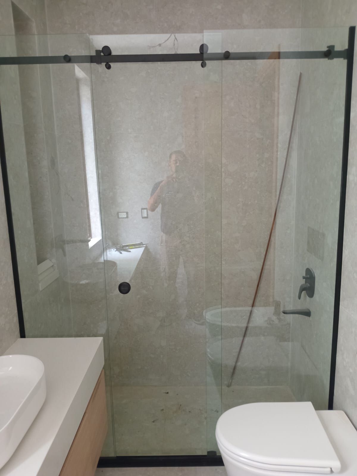 Baño con puerta de ducha de cristal, inodoro, lavabo y reflejo de una persona en el cristal.