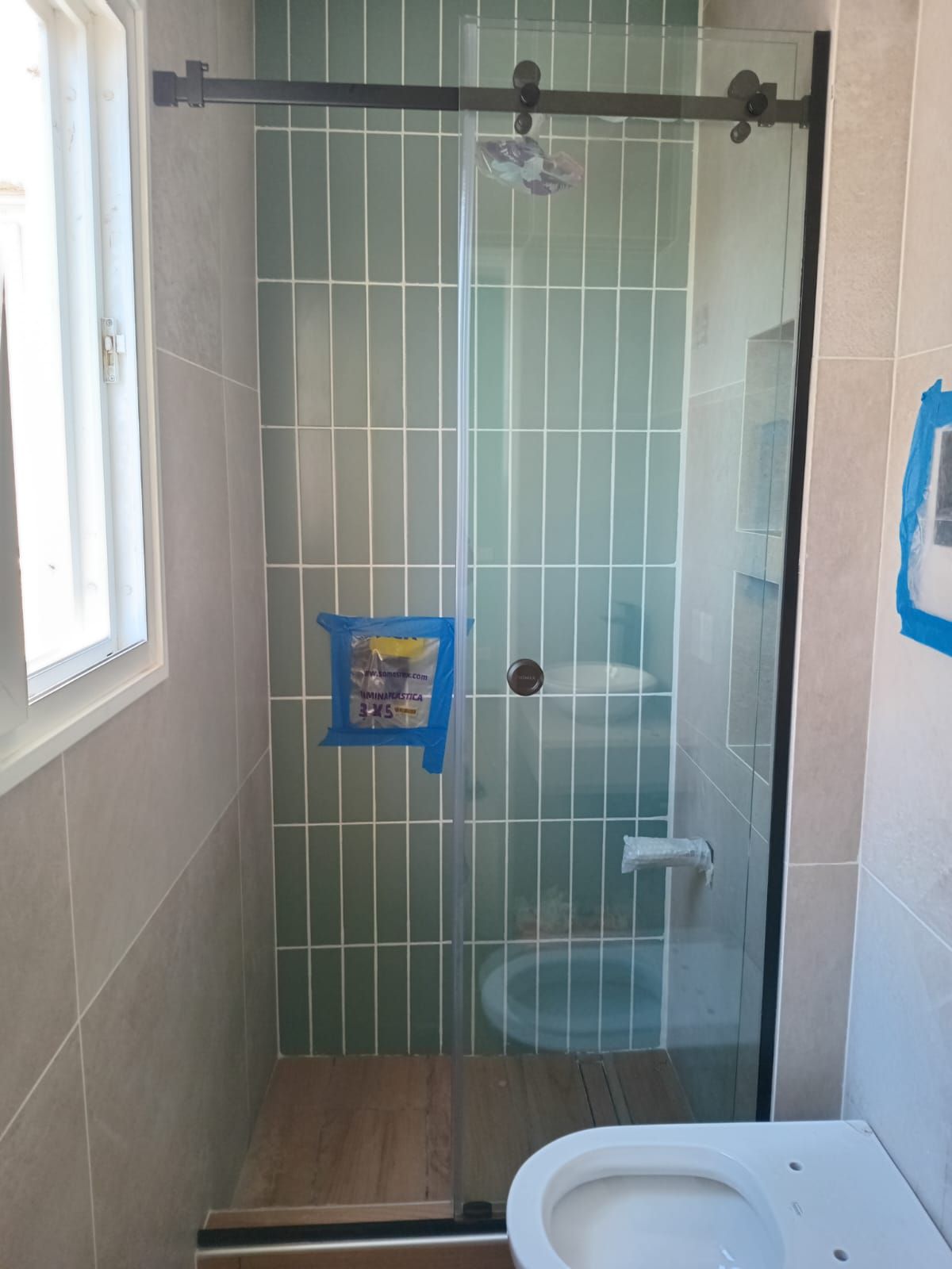 Baño con puerta de ducha de cristal, pared de azulejos verdes y inodoro blanco.