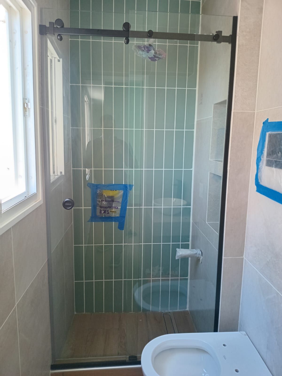 Baño con puerta de ducha de vidrio, pared de azulejos verdes, herrajes de puerta negros e inodoro.