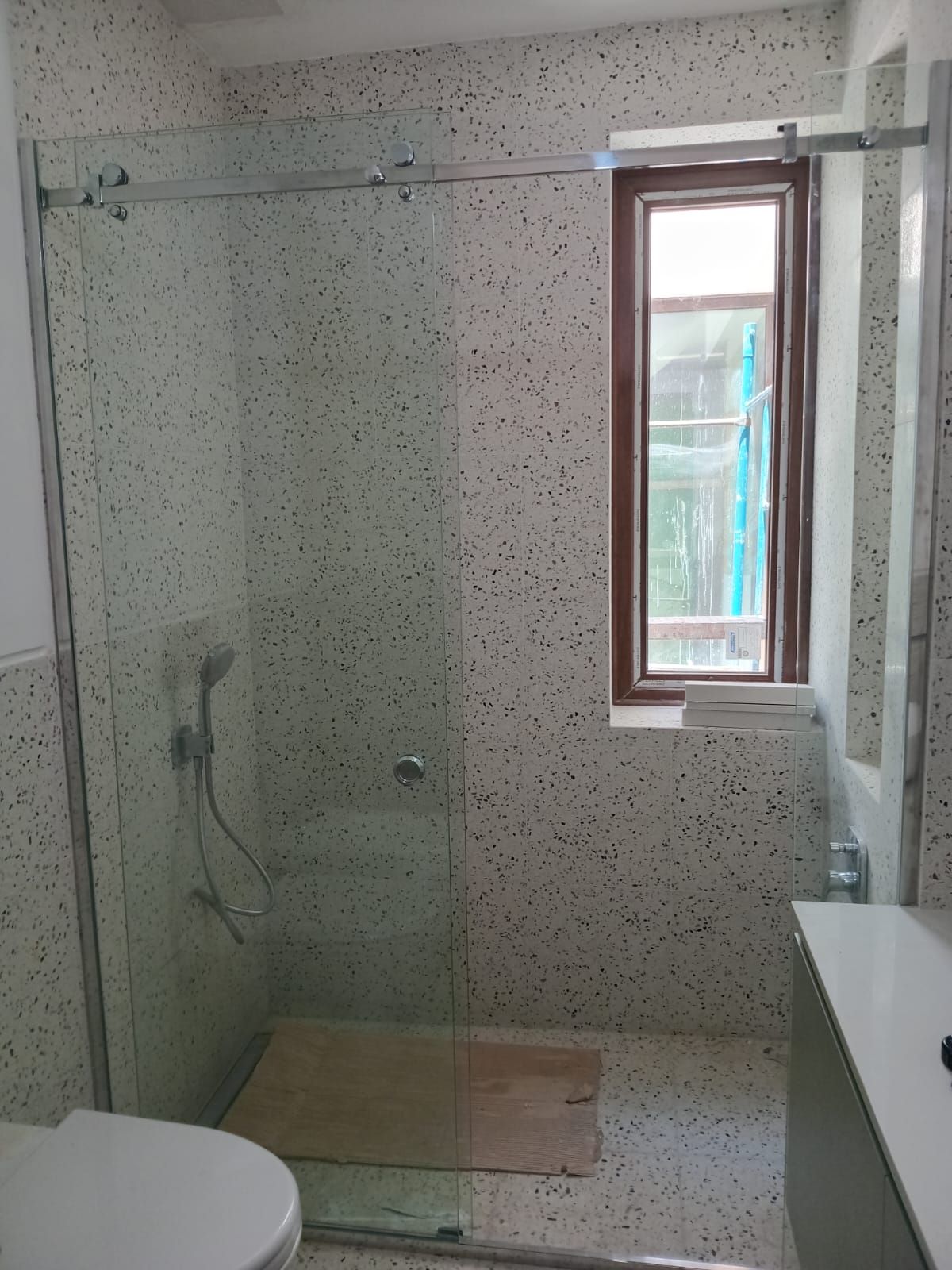 Baño con azulejos blancos moteados, puerta de ducha de cristal y una ventana con marco marrón.