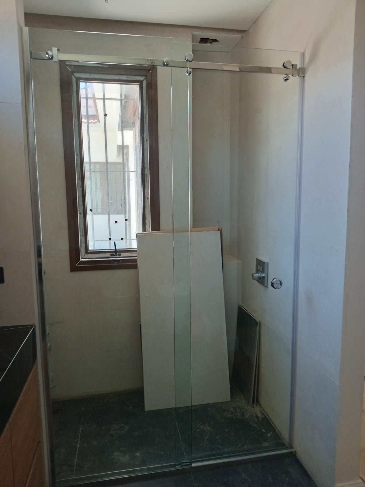 Cabina de ducha con puertas de cristal, junto a una ventana. Paredes blancas, molduras de madera. 