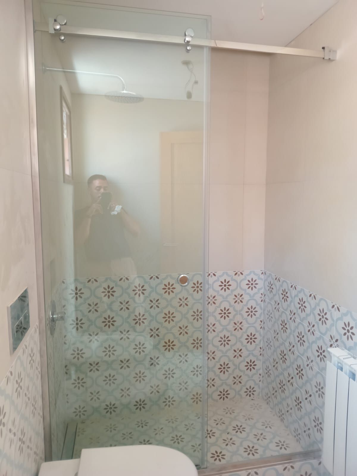 Baño con puerta de ducha de cristal, azulejos estampados y reflejo de una persona.