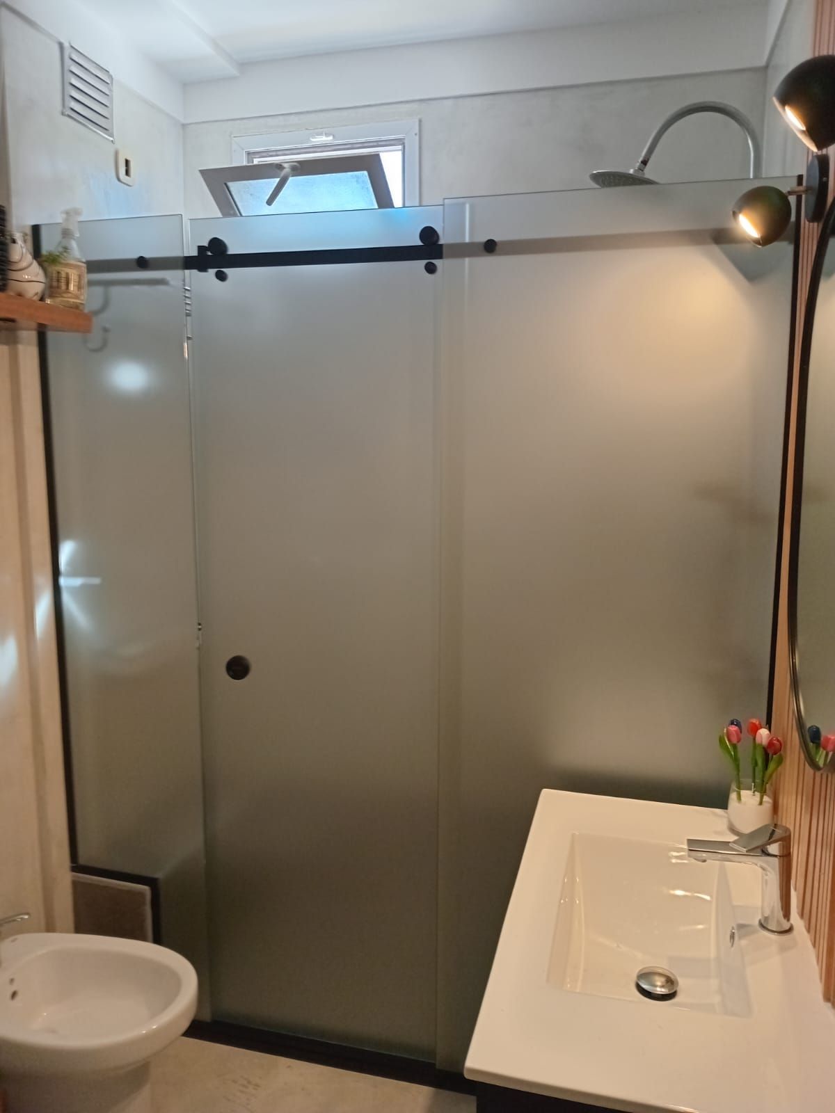 Baño con puertas de ducha de vidrio esmerilado, lavabo blanco, inodoro y pequeña ventana.