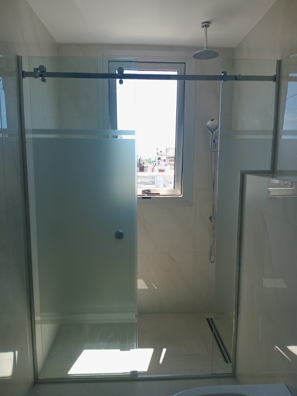 Cabina de ducha con puerta de vidrio esmerilado, adyacente a una ventana, con cabezal de ducha tipo lluvia.
