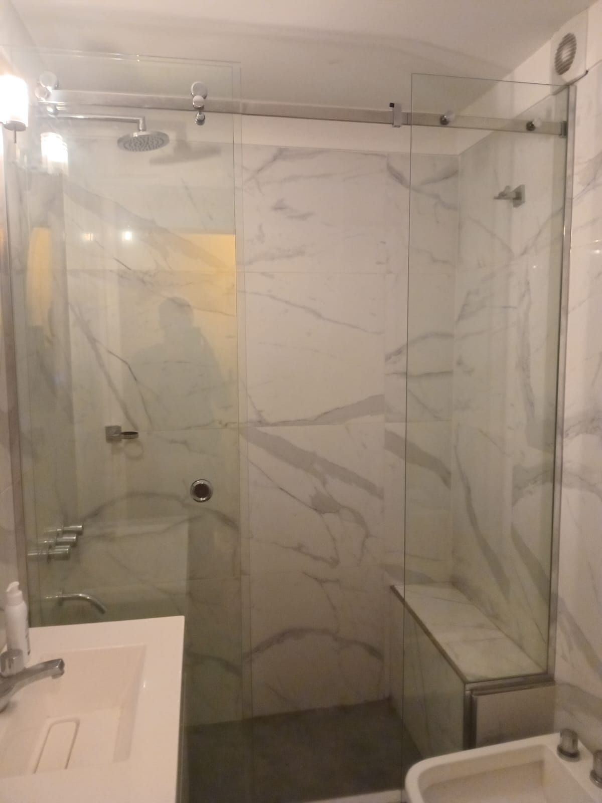 Baño con paredes de mármol y mampara de ducha de cristal. Lavabo e inodoro blancos visibles.