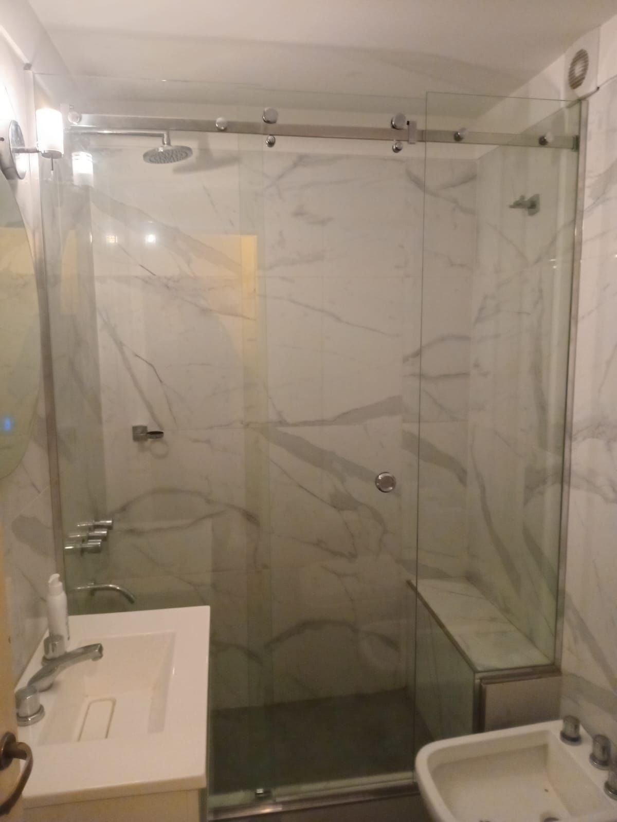 Baño con revestimiento de pared efecto mármol, mampara de ducha de cristal con puerta corredera y lavabo blanco.
