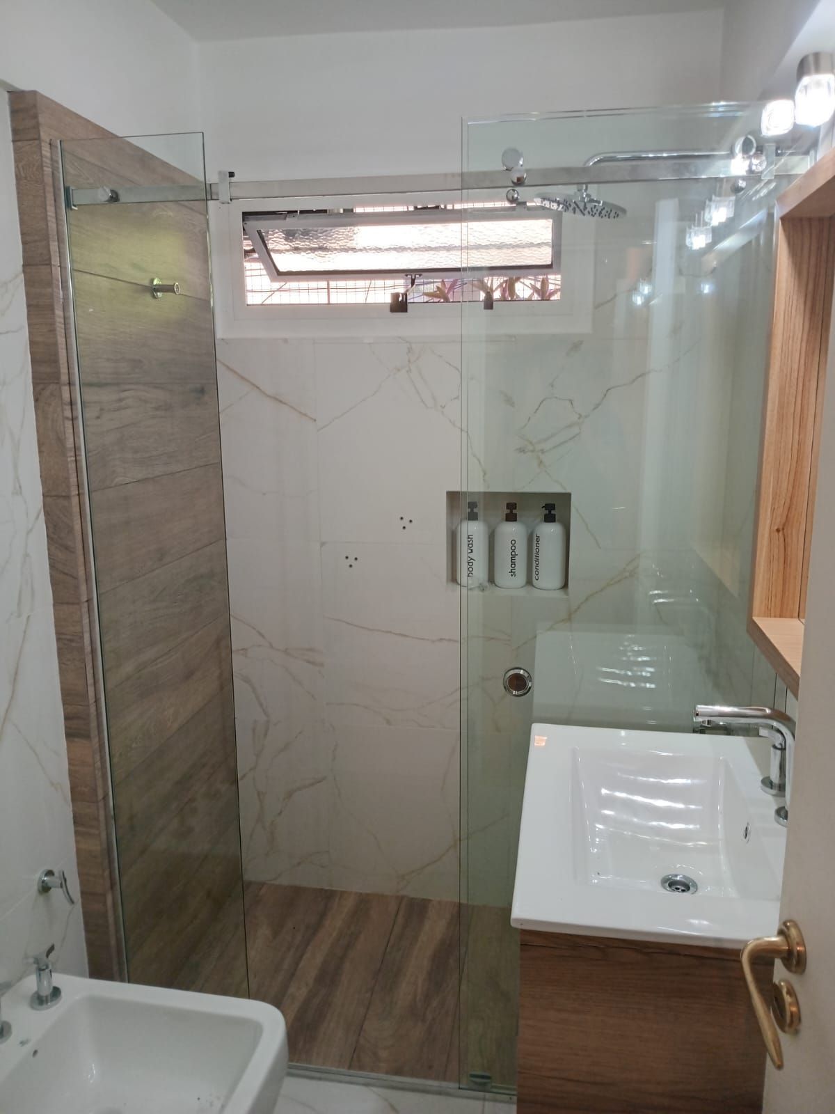 Baño moderno con ducha de cristal, lavabo blanco y azulejos con aspecto de madera.