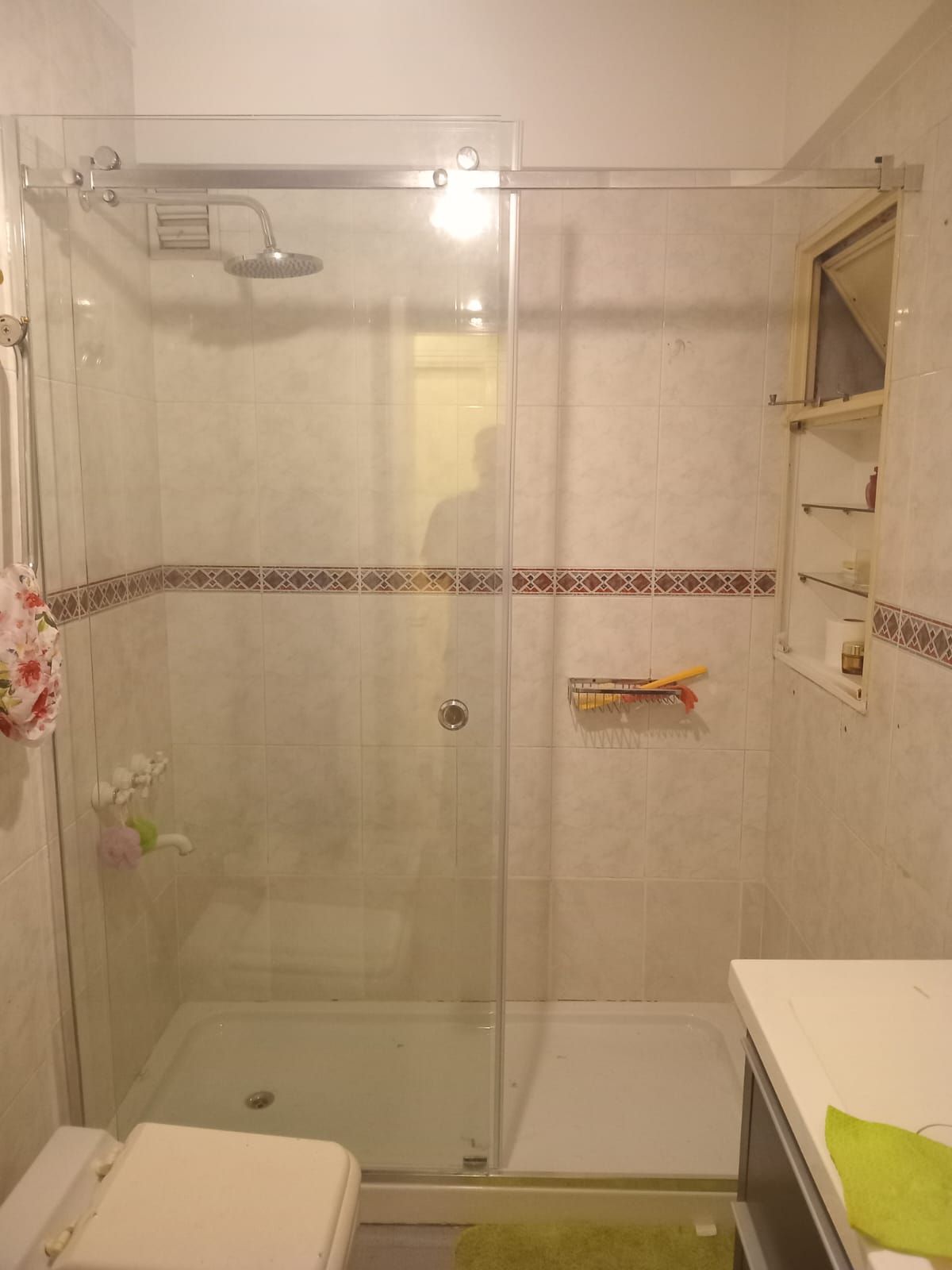 Ducha con mampara de cristal y paredes de azulejos blancos. Inodoro a la izquierda, lavabo y trastero a la derecha.