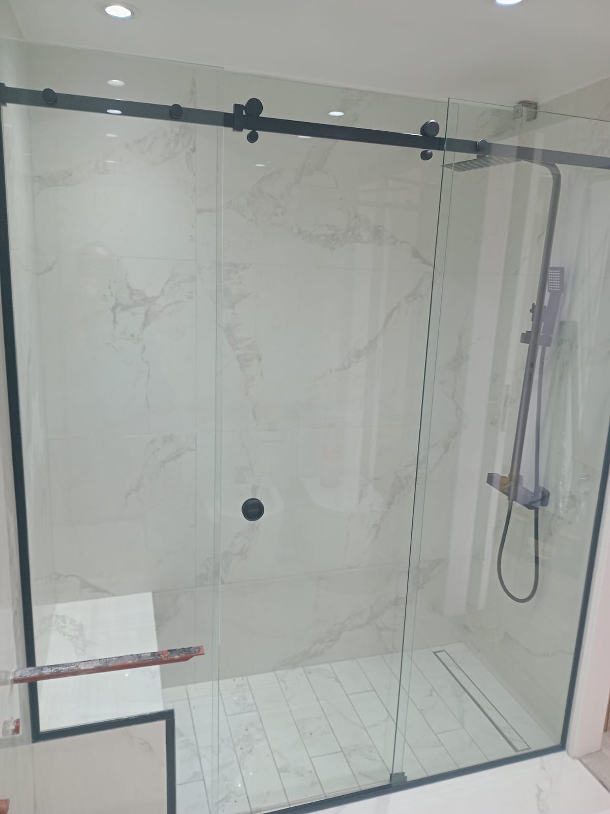 Mampara de ducha con moldura negra, puerta corrediza de cristal, azulejos blancos y cabezal de ducha.