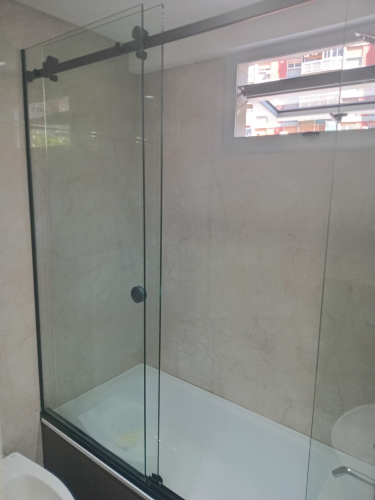 Puerta de ducha de vidrio con herrajes negros instalada sobre una bañera. Paredes de azulejos beige y ventana.