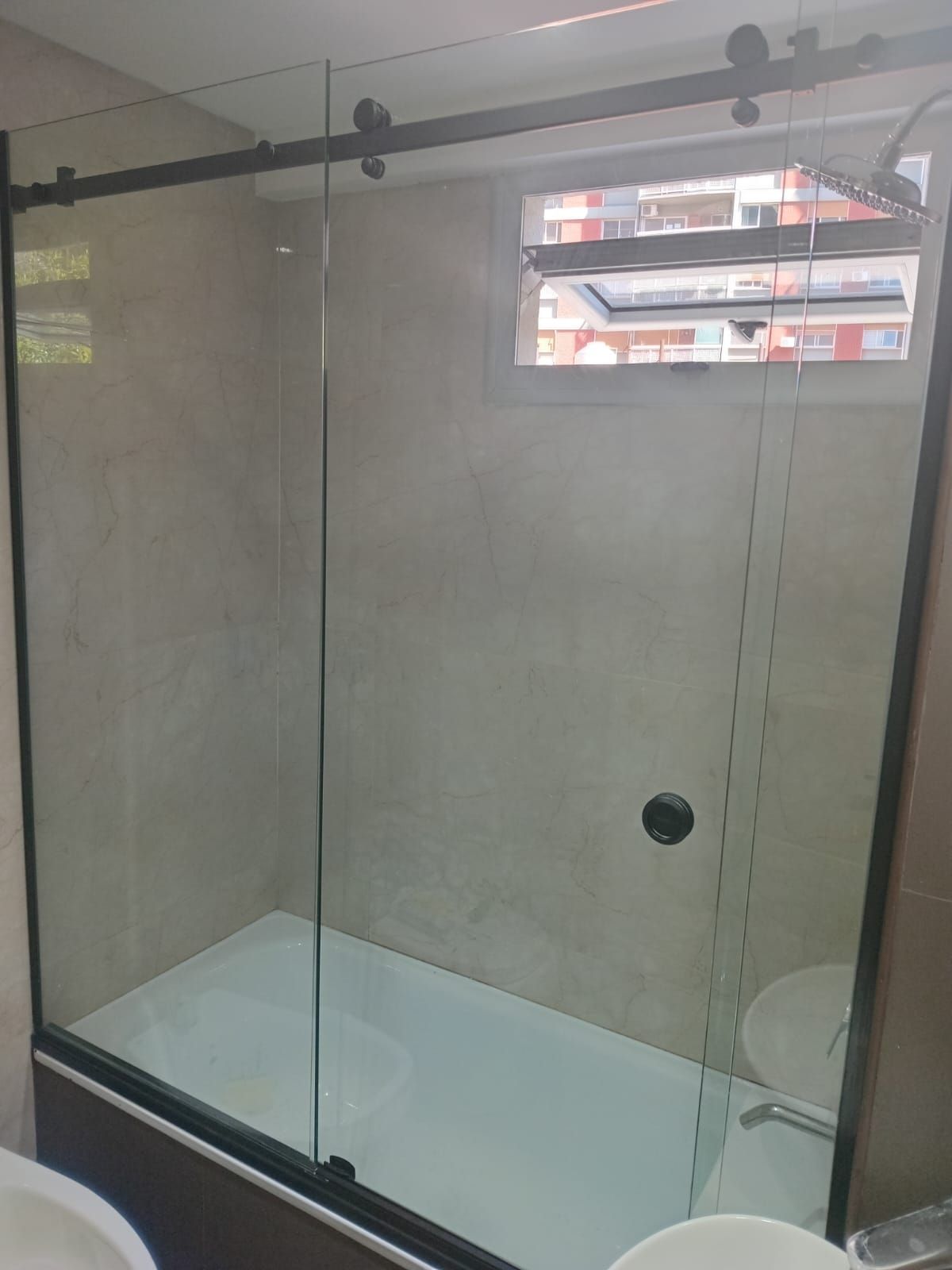 Mampara de ducha con puerta corredera de cristal encima de una bañera, al lado de una ventana.