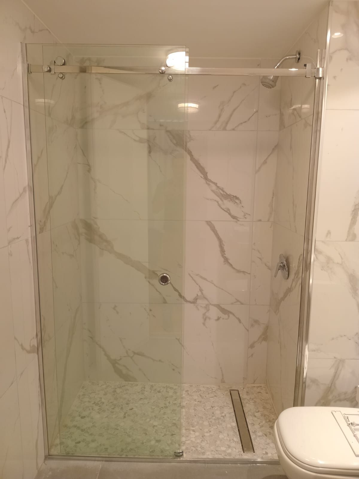 Cabina de ducha con puerta corrediza de cristal y azulejos imitación mármol. Grifería de acero inoxidable.