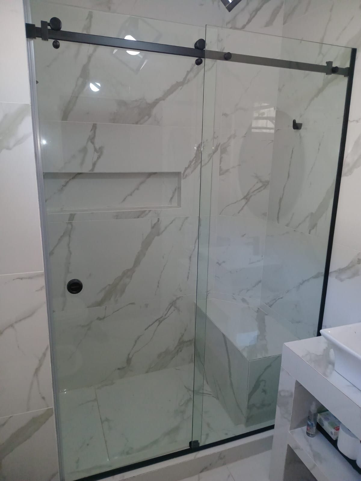 Mampara de ducha de cristal con herrajes negros, azulejos con estampado de mármol y banco incorporado.