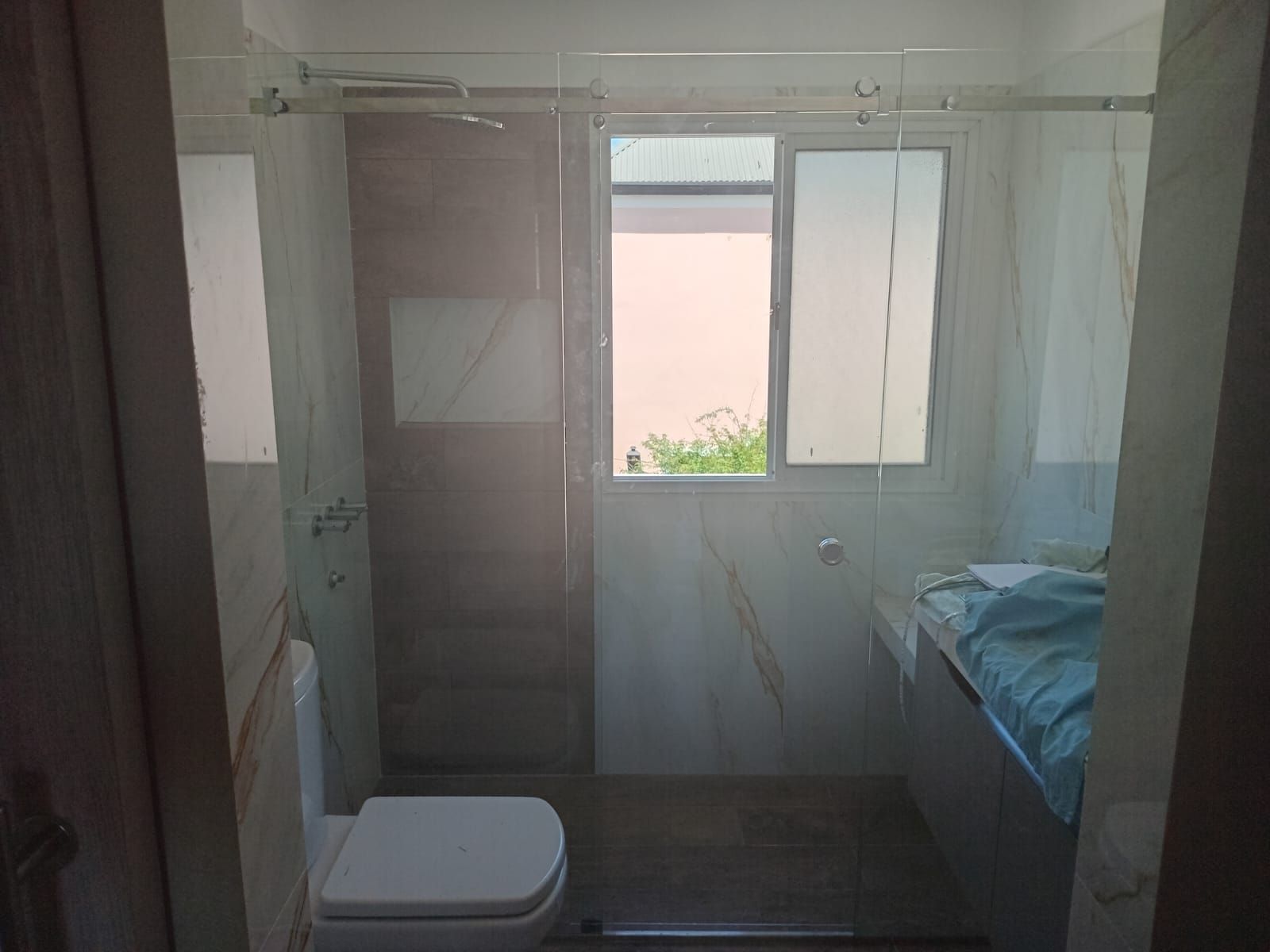Baño con inodoro, ventana y ducha. Azulejos claros y mampara de cristal.
