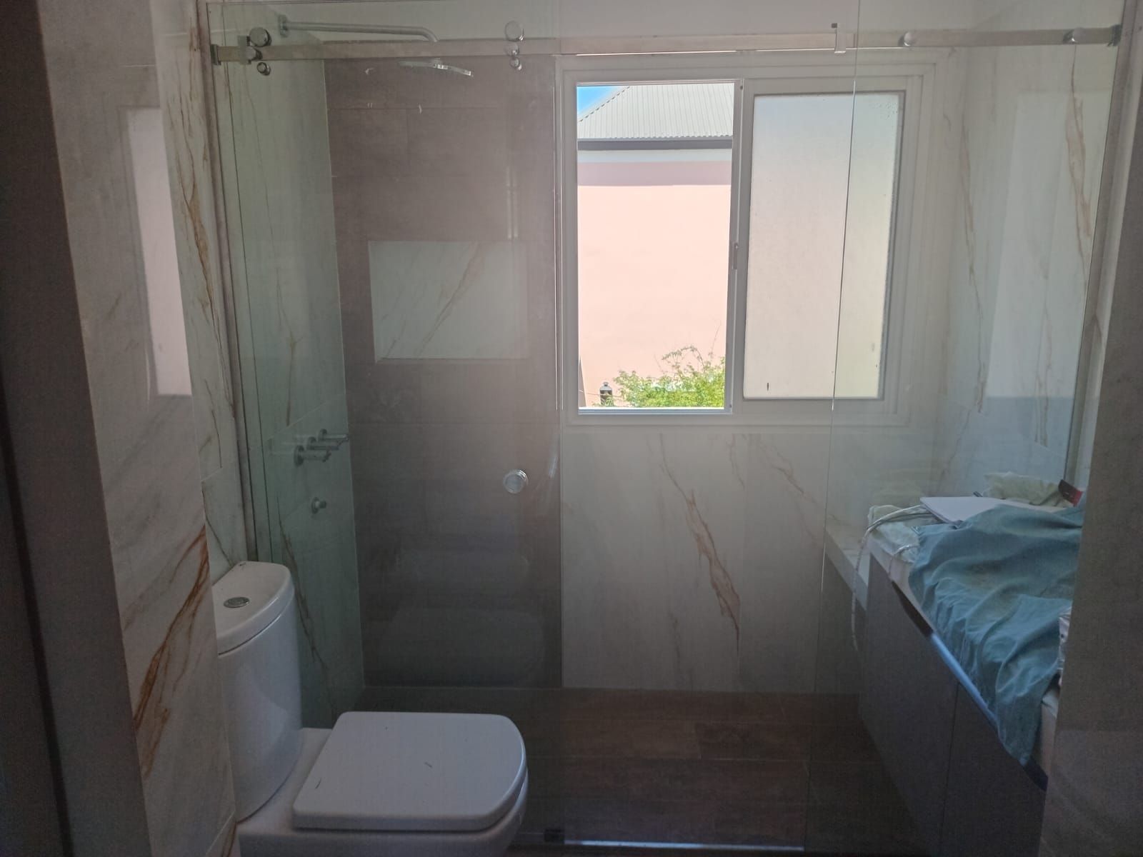 Baño con ducha de cristal, inodoro, ventana de cristal esmerilado y tocador. Paredes con efecto mármol.