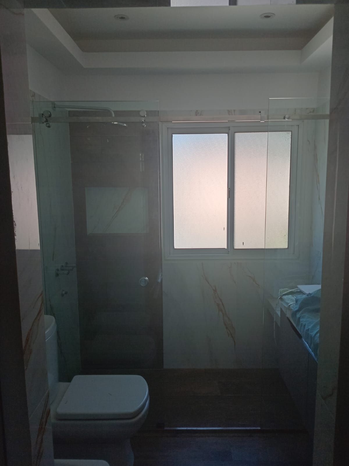 Interior de baño con mampara de ducha de cristal, ventana esmerilada, inodoro y tocador.
