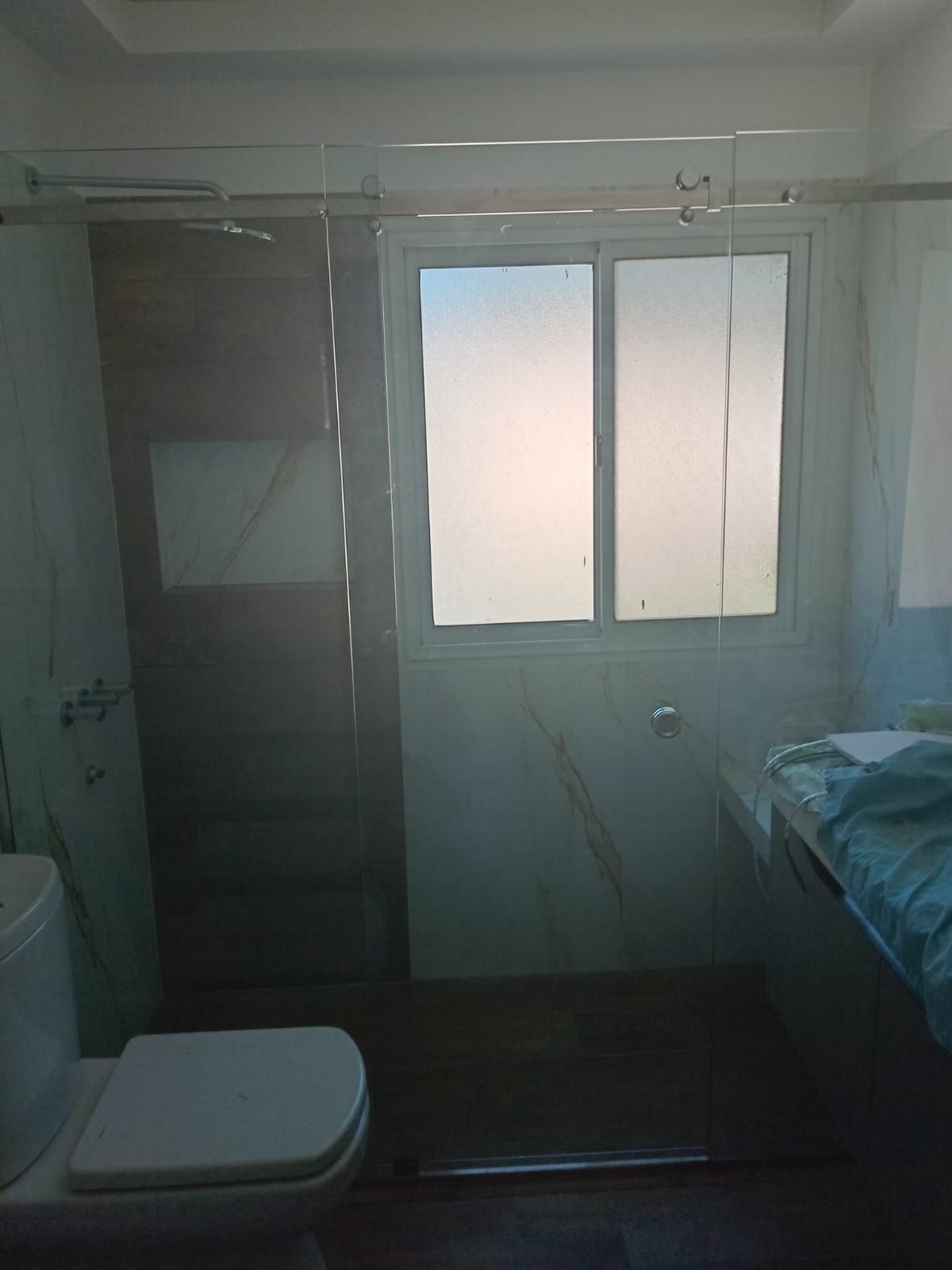 Baño con inodoro, ducha y ventana. Paredes de azulejos blancos y mármol, y toalla azul.