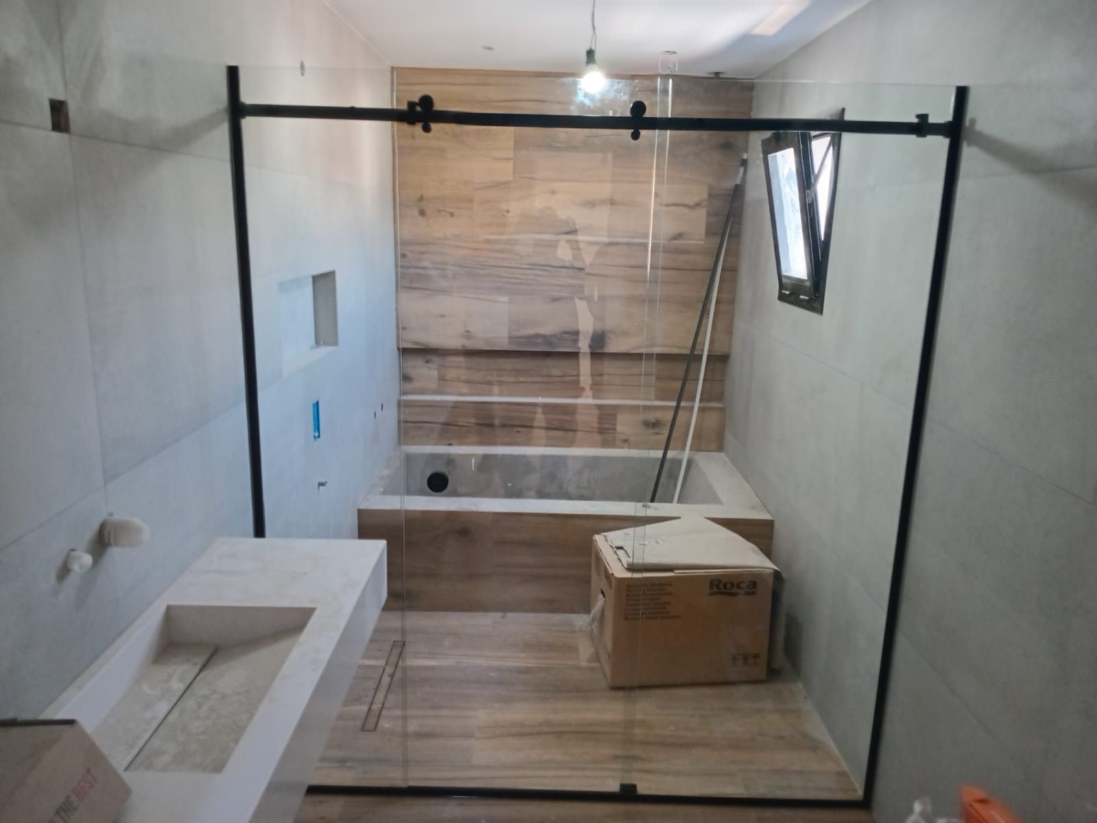 Baño en construcción con mampara de ducha de cristal, azulejos imitación madera y tocador moderno.