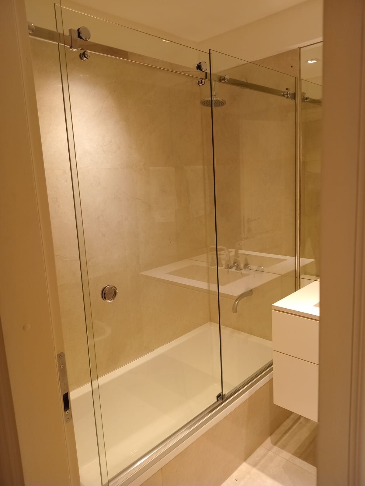 Mampara de ducha de cristal sobre bañera con puerta corredera. Azulejos beige y grifería moderna.