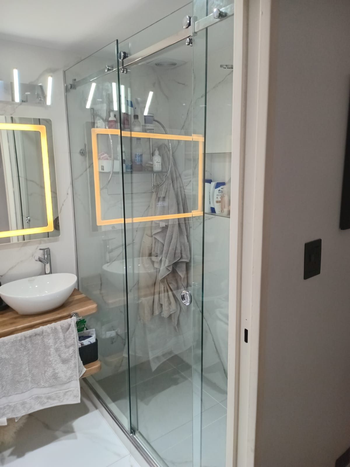 Baño moderno con mampara de ducha de cristal, tocador con lavabo sobre encimera y espejo iluminado.