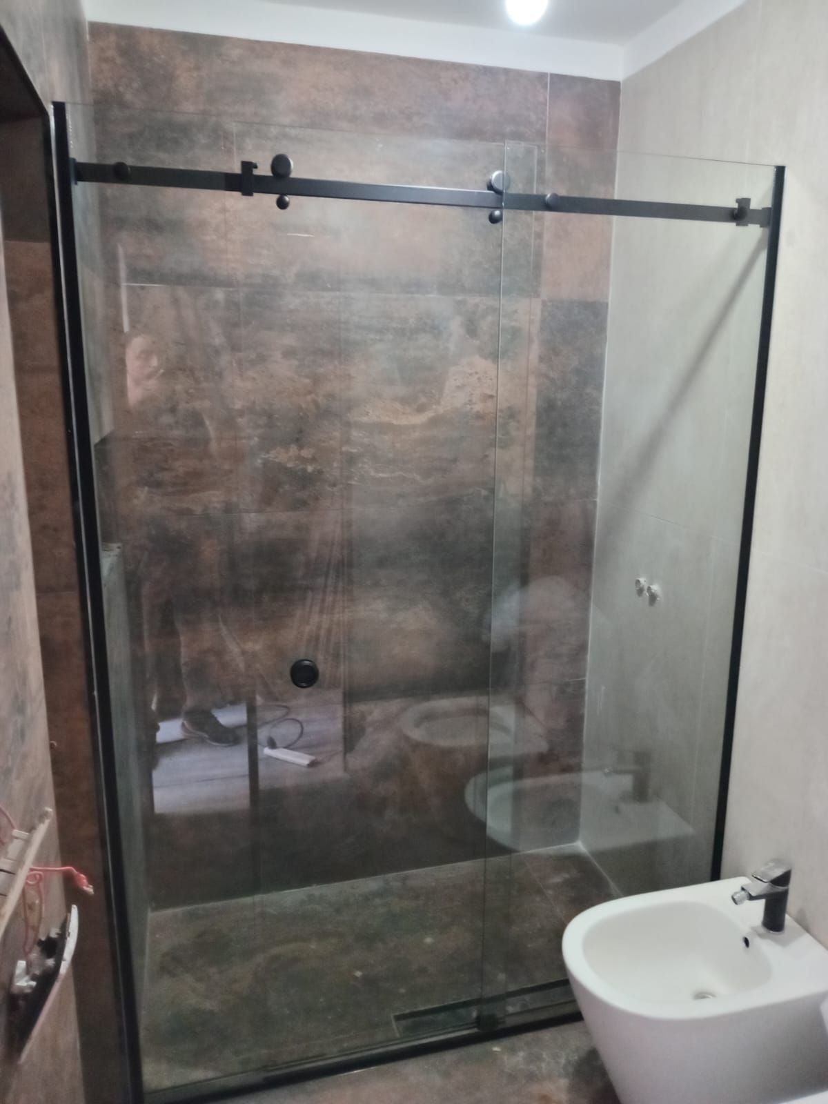 Mampara de ducha corrediza de cristal con marco negro en un baño con azulejos marrones y blancos y bidé.