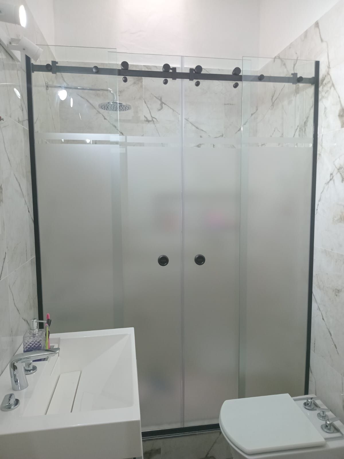 Mampara de ducha de vidrio esmerilado con marco negro en un baño. Lavabo e inodoro blancos visibles.