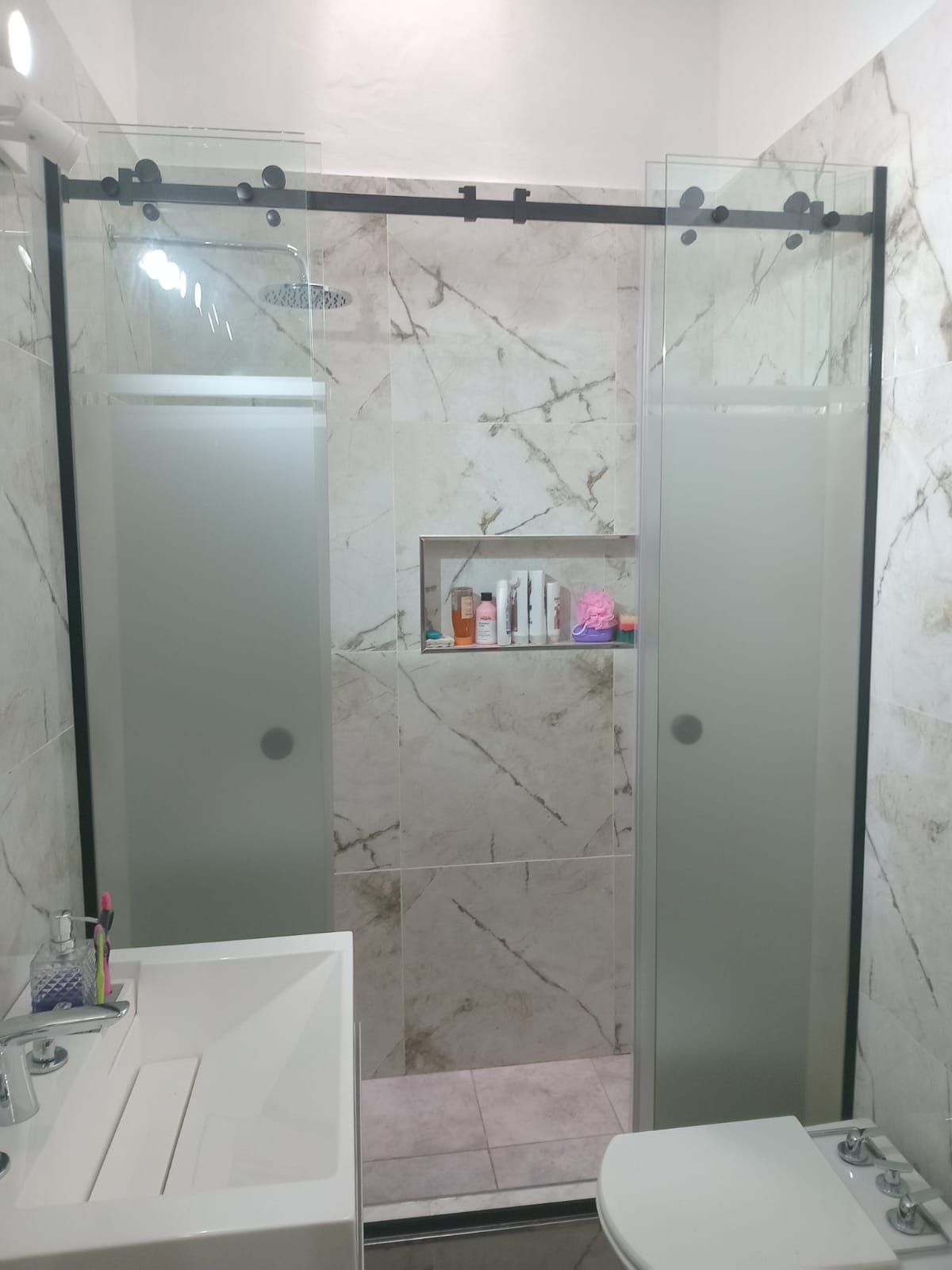 Baño con puertas de ducha corredizas de vidrio, marco negro y pared de azulejos con diseño de mármol.