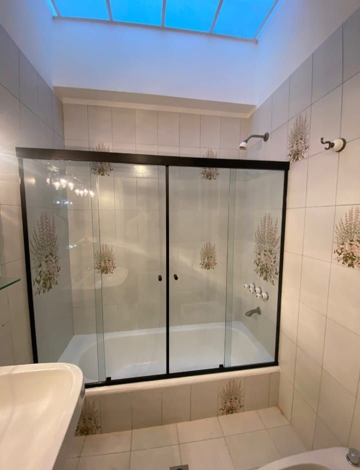 Baño con mampara de cristal sobre la bañera. Azulejos blancos y beige, y claraboya en la parte superior.