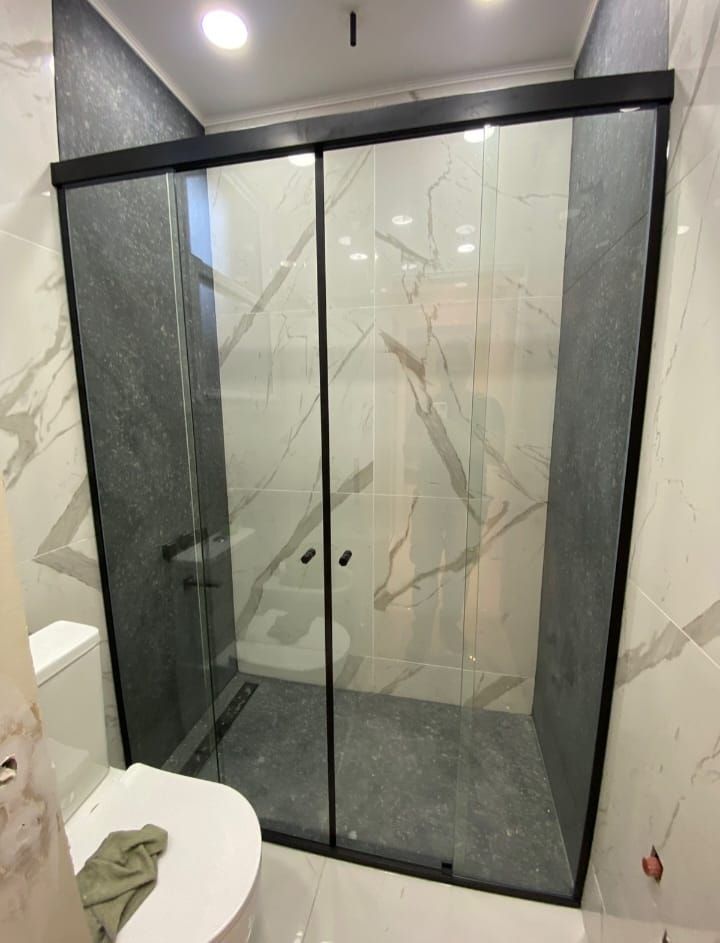 Mampara de ducha de cristal con marco negro en un baño con paredes de mármol, inodoro a la vista.