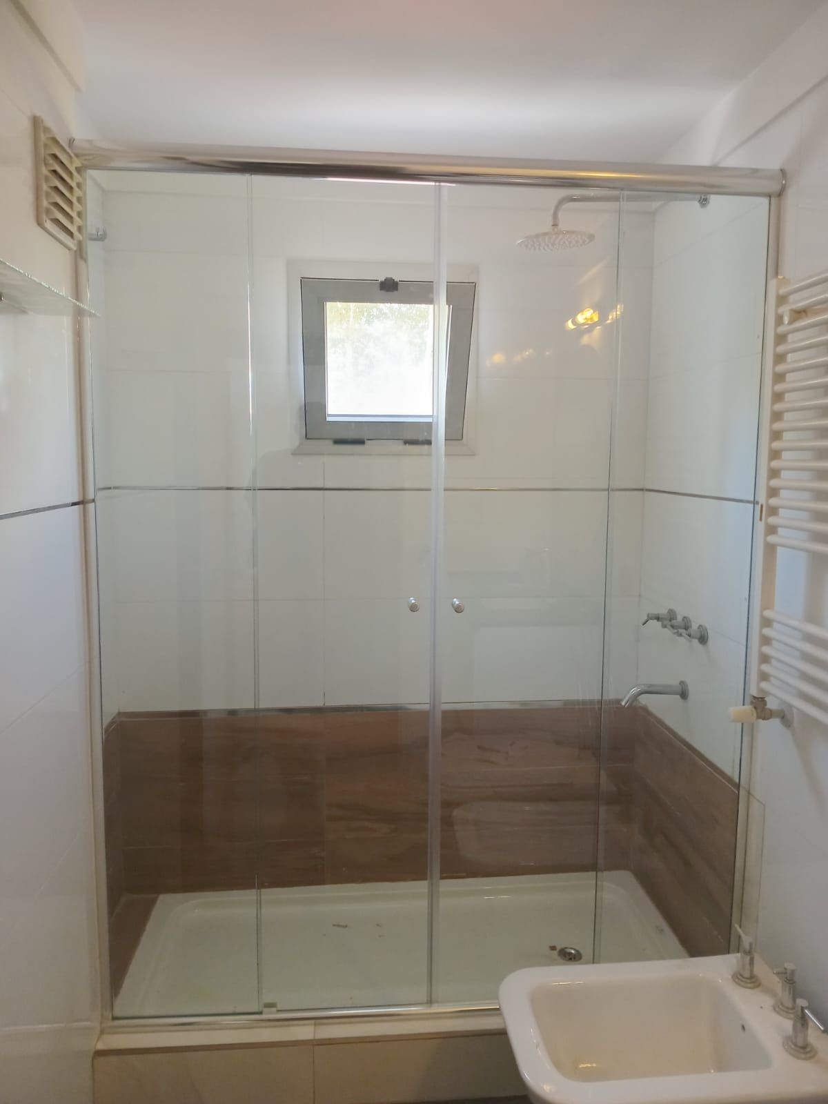 Cabina de ducha con puertas corredizas de vidrio, pequeña ventana y paredes de azulejos blancos.