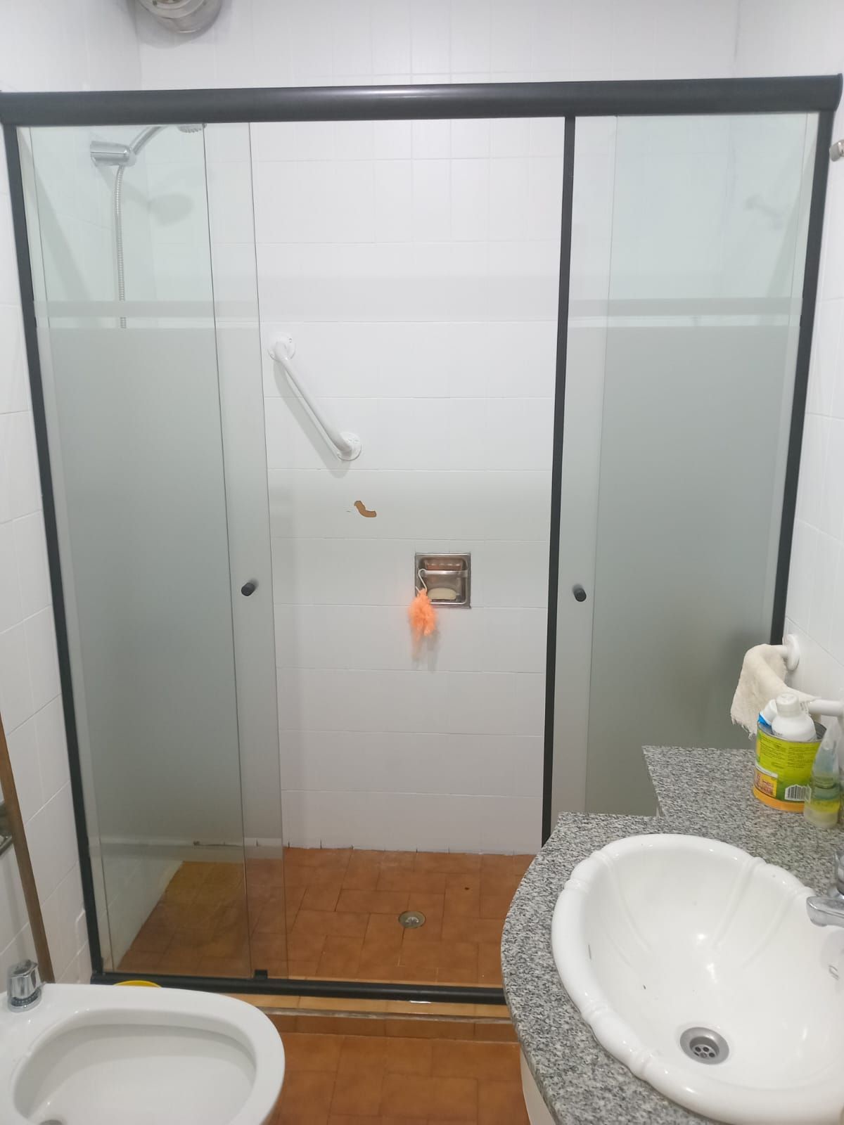 Baño con puerta de ducha de cristal, paredes blancas, lavabo, inodoro y suelo marrón.