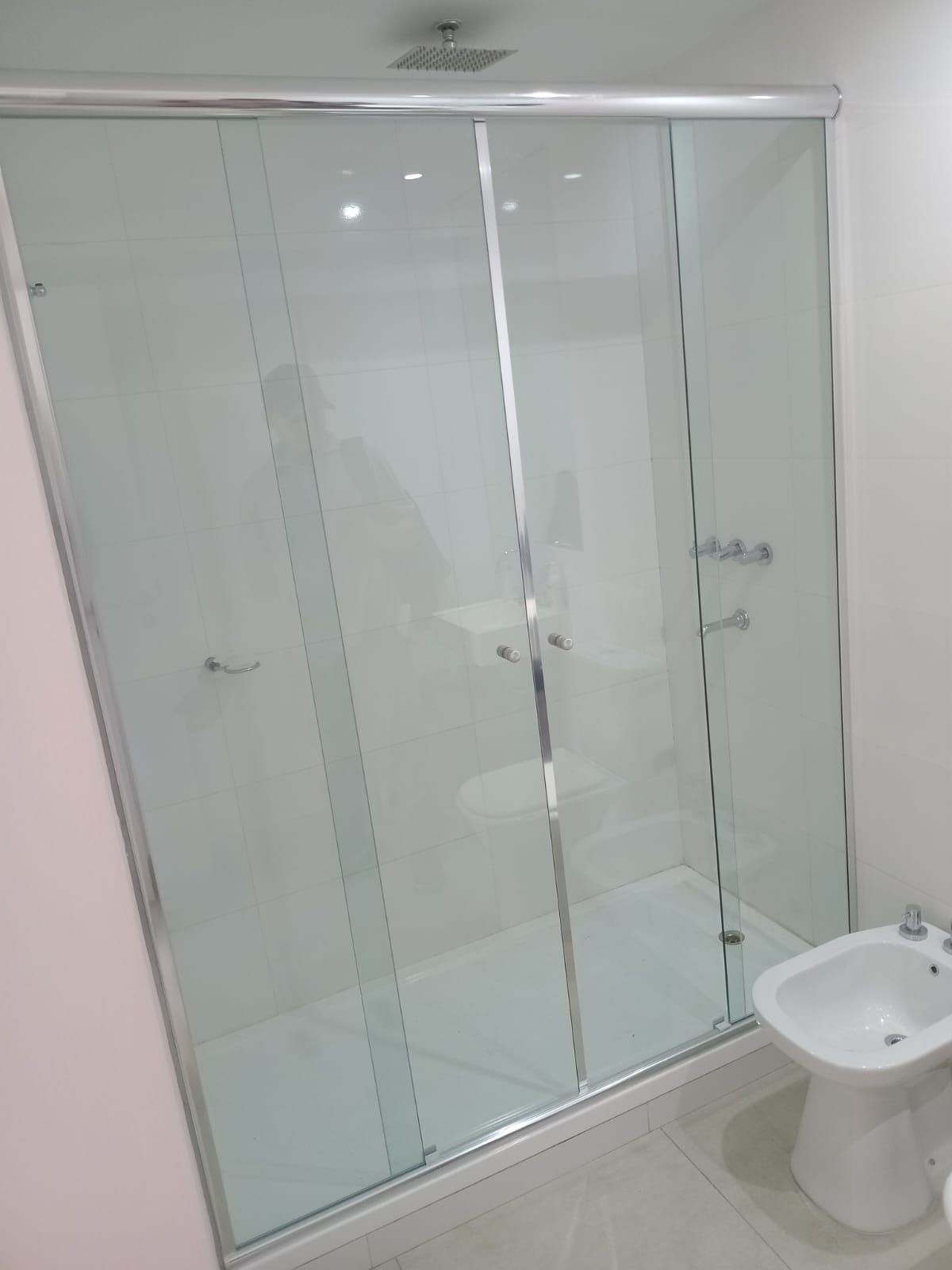 Cabina de ducha con puertas corredizas de vidrio, molduras cromadas y bidé en un baño blanco.