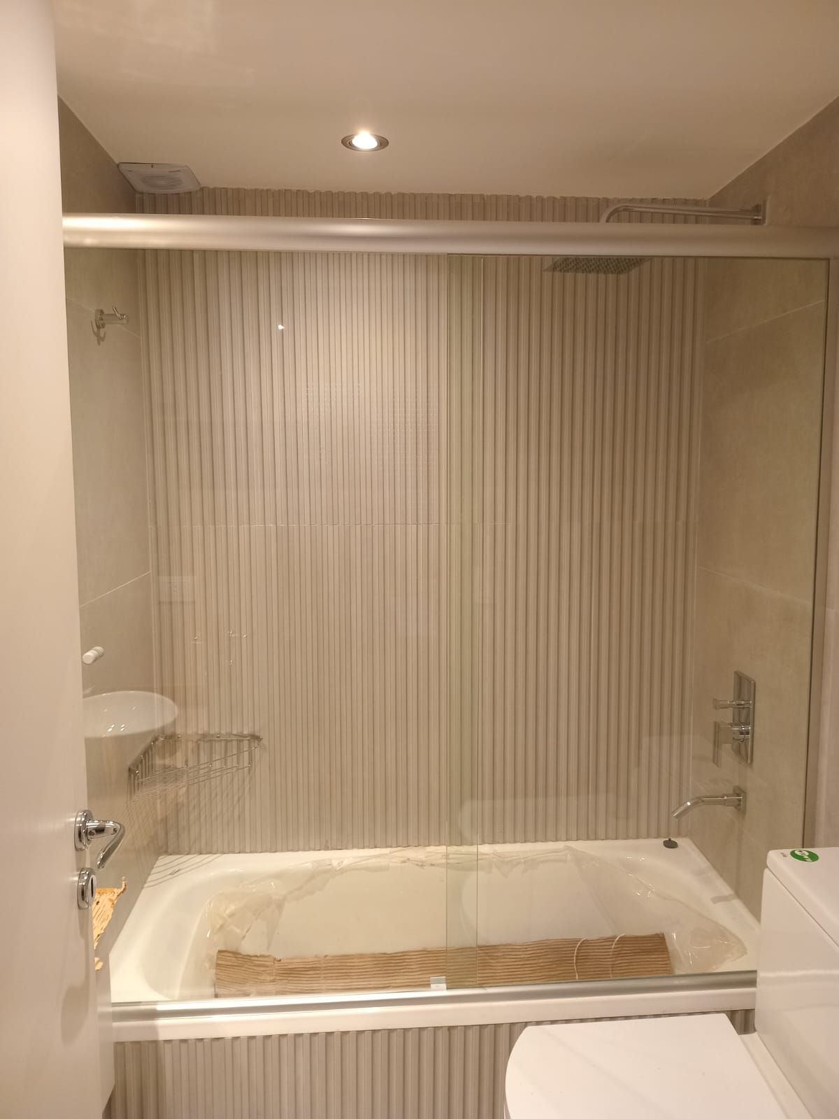 Baño con bañera blanca, mampara de cristal y revestimiento de pared con rayas verticales.
