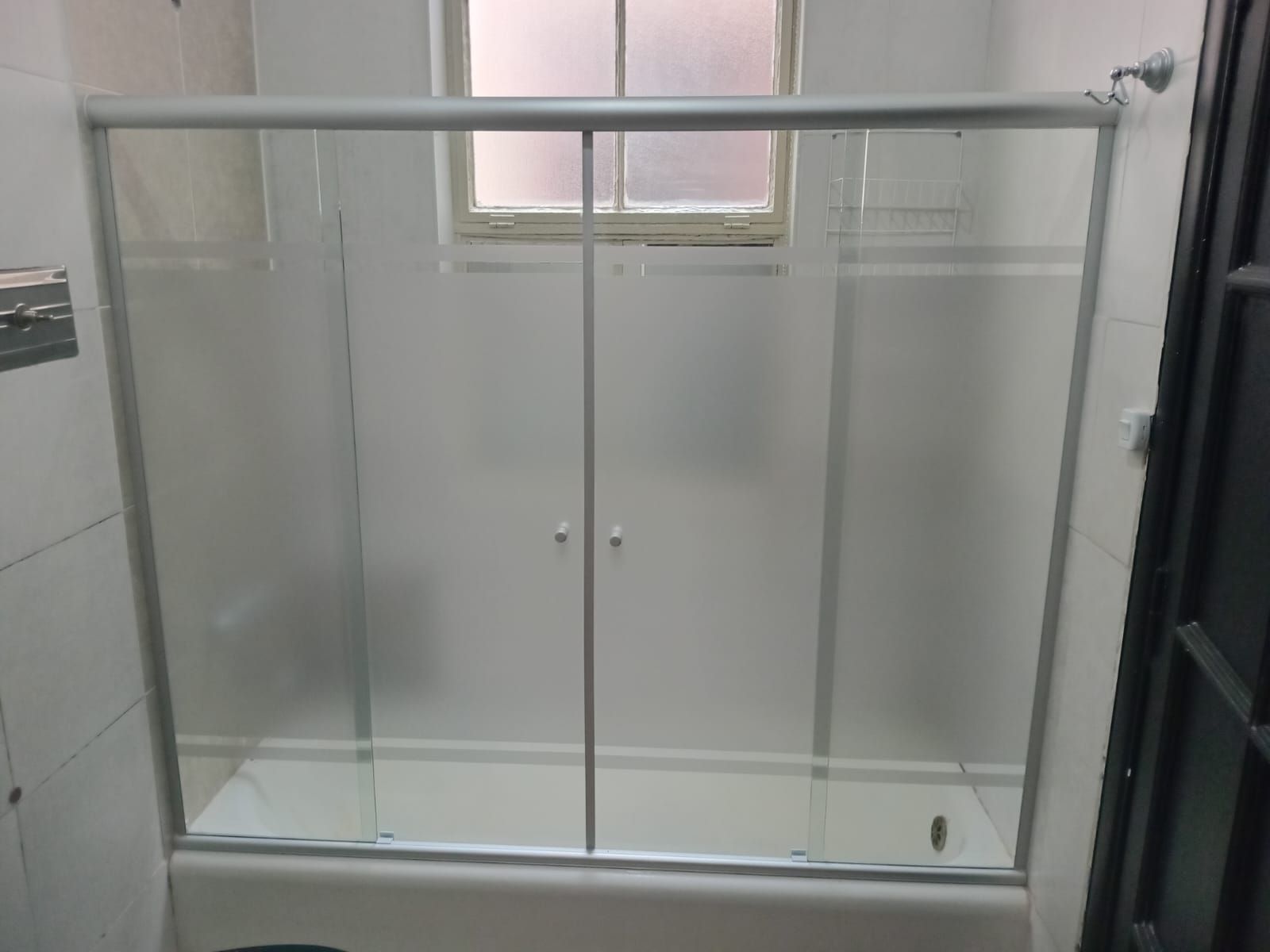 Cabina de ducha con puertas de vidrio esmerilado y marco de metal, sobre azulejos blancos.