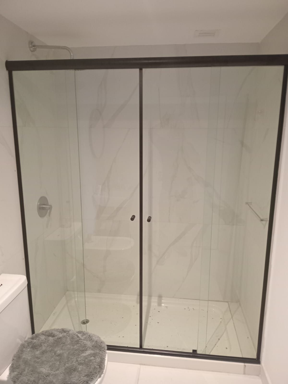 Cabina de ducha con puertas corredizas de vidrio, molduras oscuras y azulejos de pared con apariencia de mármol.