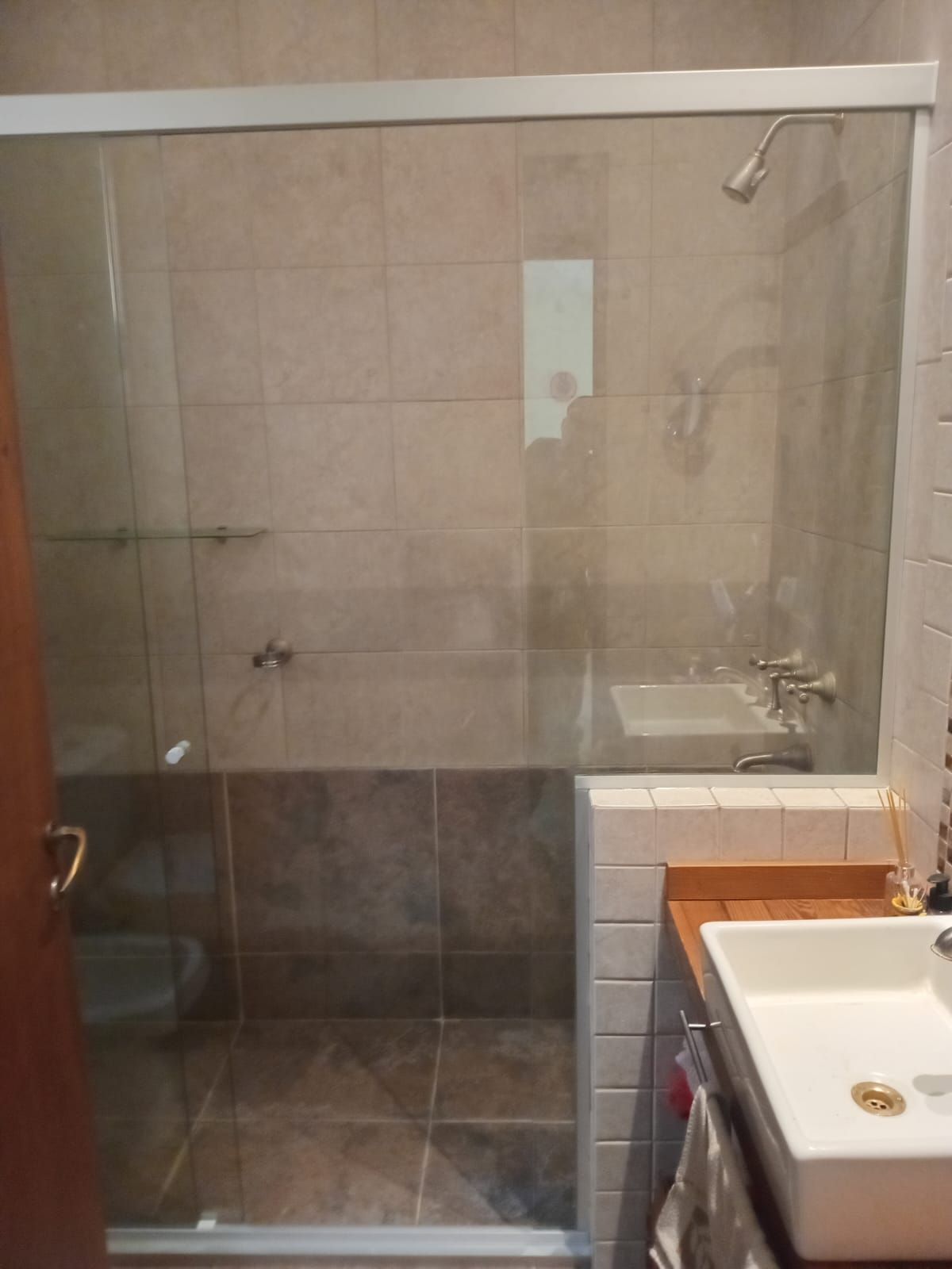Ducha con puerta de cristal, azulejos beige, accesorios plateados y lavabo blanco sobre encimera de madera.