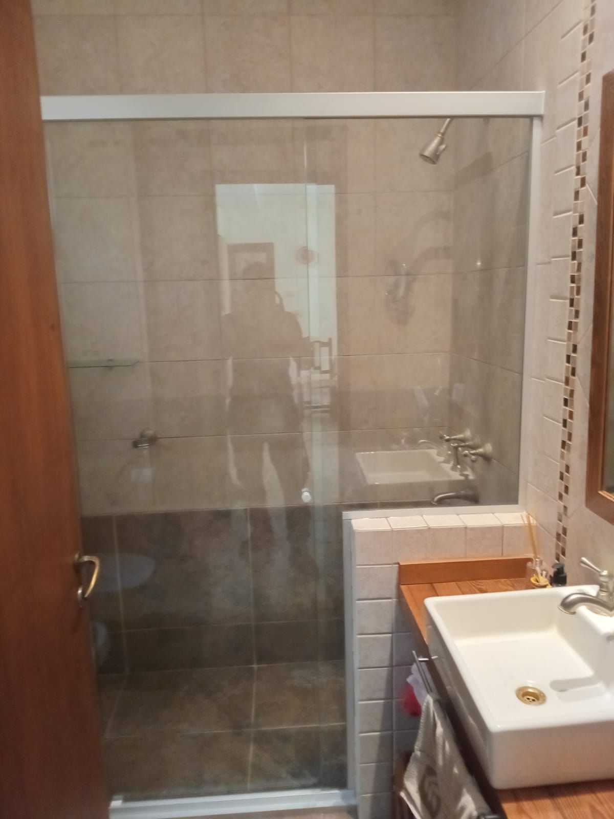 Baño con ducha de cristal, lavabo e inodoro. Azulejos beige y puerta de madera.
