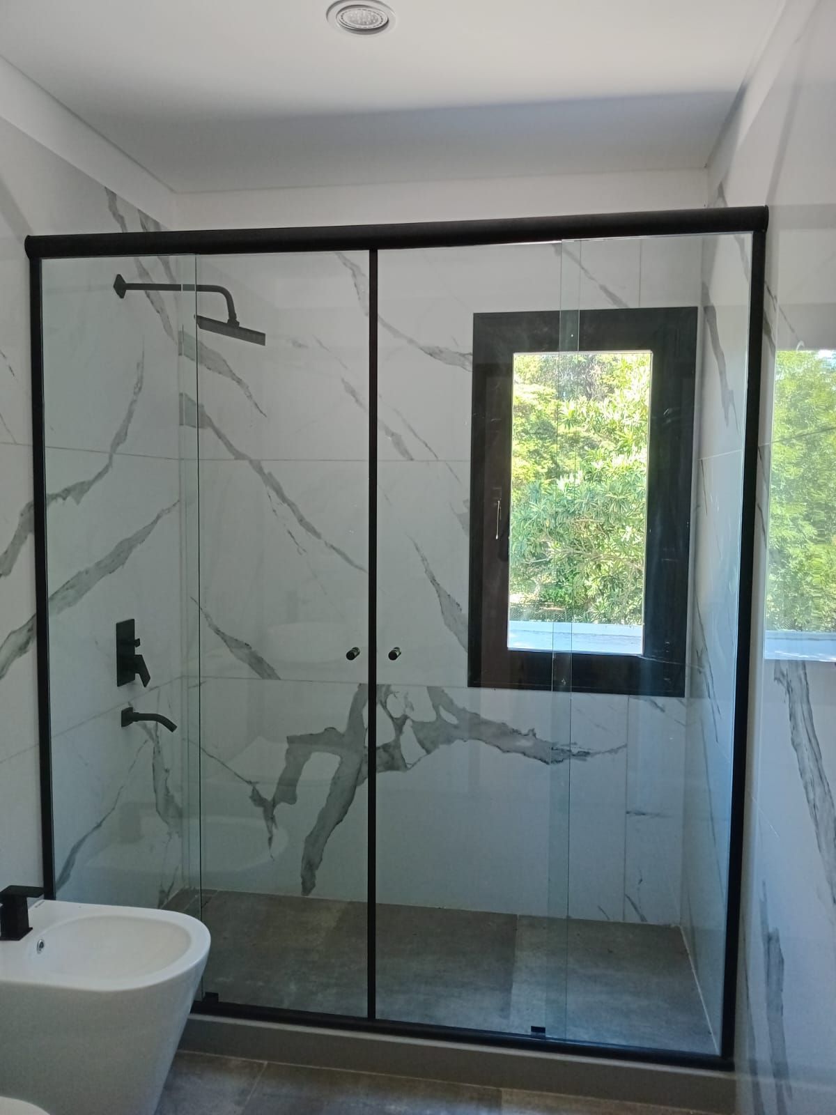 Baño moderno con azulejos de mármol, ducha con marco negro con puertas de vidrio y ventana.