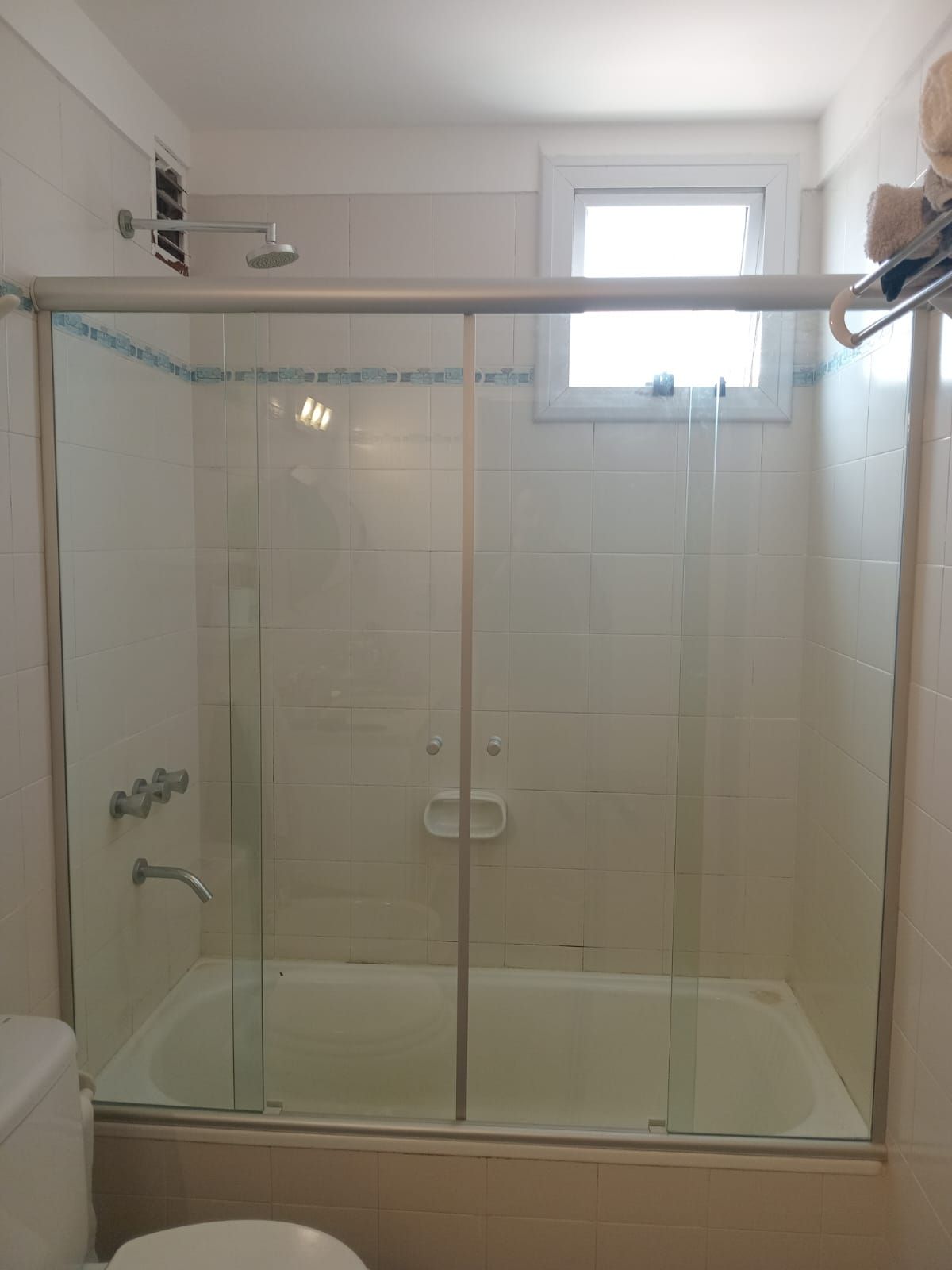 Mampara de ducha/bañera con puertas corredizas de cristal, azulejos blancos, una pequeña ventana y un cabezal de ducha.