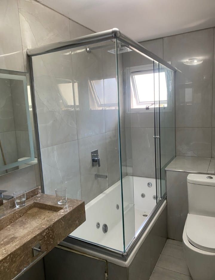 Baño con cabina de ducha de cristal sobre una bañera blanca con chorros, lavabo de piedra beige y un inodoro blanco.