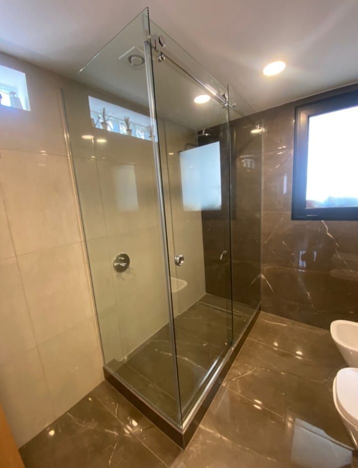 Ducha con mampara de cristal en un baño moderno con suelo y paredes de baldosas marrones y una pequeña ventana.