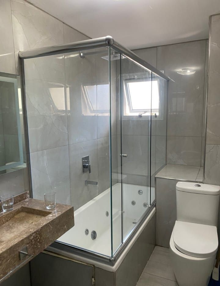 Baño con mampara de ducha de cristal sobre bañera de hidromasaje, combinación de colores neutros y un inodoro.