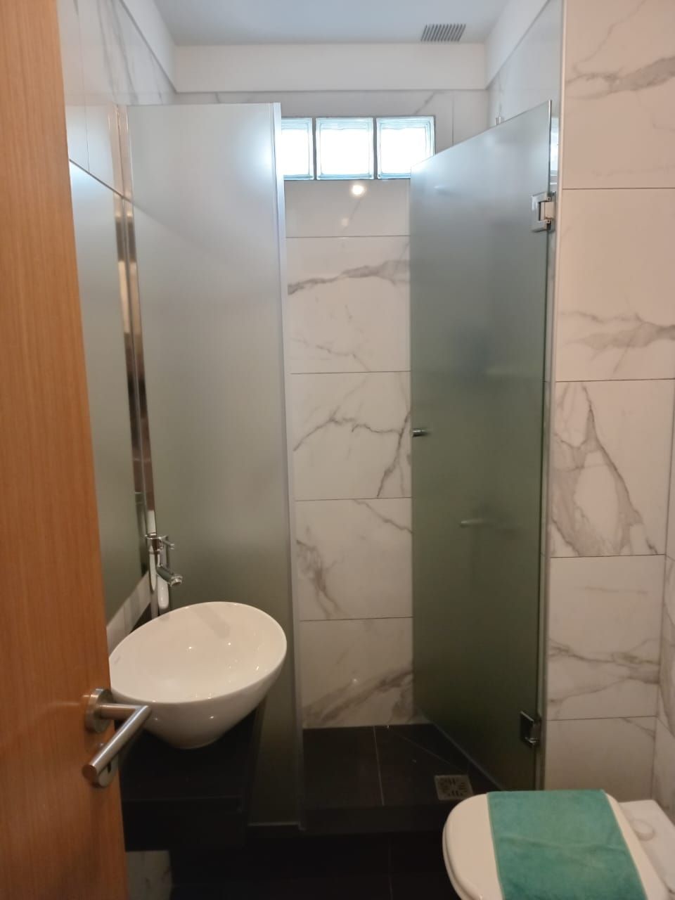 Baño con ducha, inodoro y lavabo. Azulejos de mármol blanco, suelo oscuro y mampara de cristal.