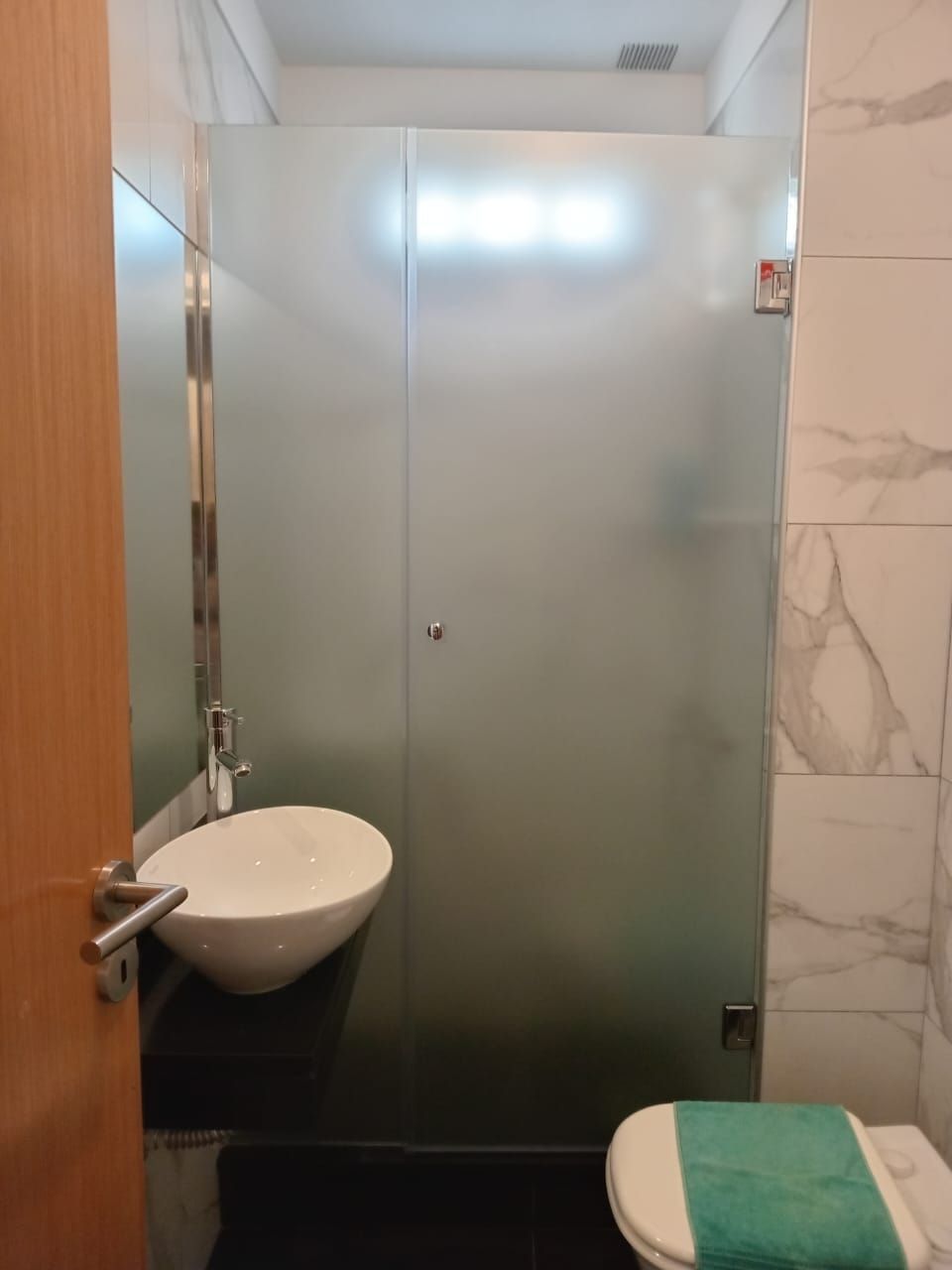 Baño con puerta de ducha de vidrio esmerilado, lavabo blanco, inodoro y pared de azulejos de mármol.